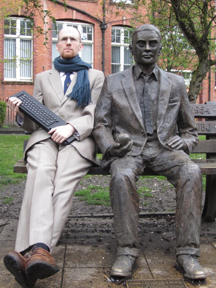 A park bench with two white men in formal dress sitting on it: one tall, thin bespectacled and bearded, holding an old computer like it's an electric guitar (Sir Clive Sinclair); the other a bronze statue (Dr Alan Turing)