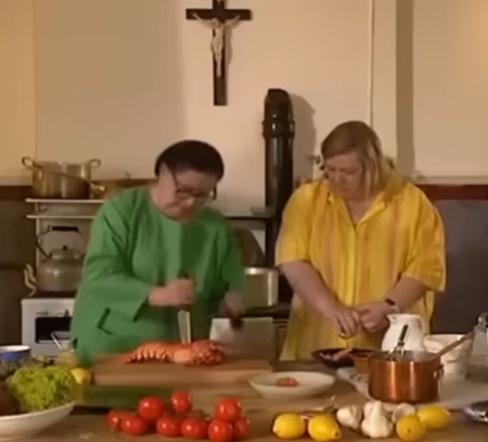 A screenshot from the British TV show Two Fat Ladies. A woman in green stabs a cooked lobster with a chef's knife beneath a crucifix while her co-host, in yellow, looks on encouraging her. 