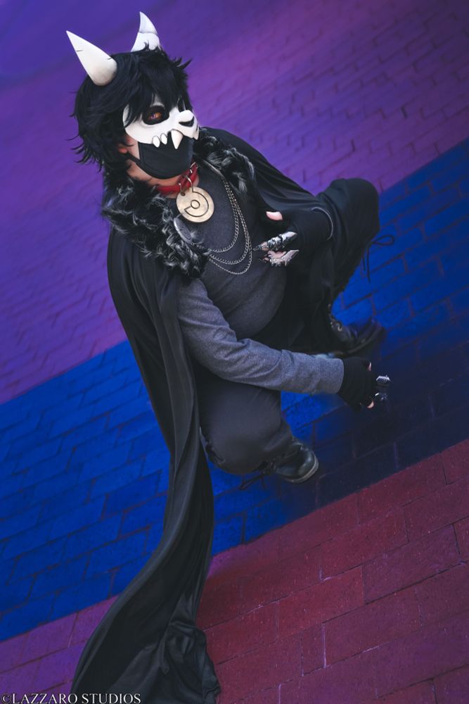Shibalove Cosplay poses as King Clawthorne from The Owl House, squatting and looking at the camera with a dark blue and maroon brick floor background 