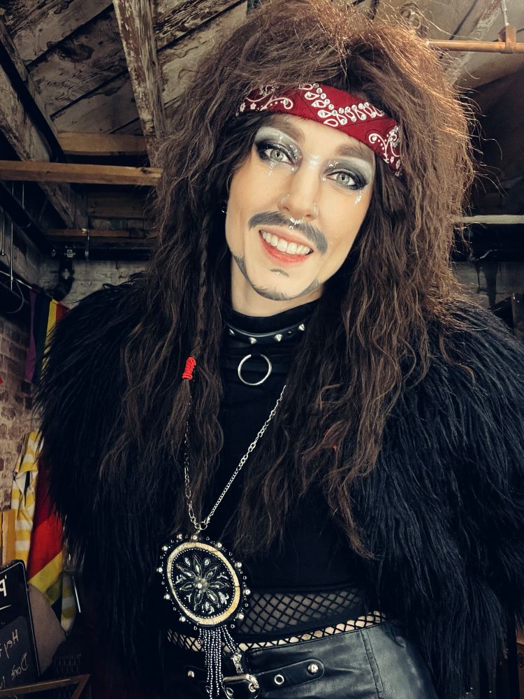 A drag performer with a mustache, beaded Métis pendant and teased 80s glam rock hairstyle smiling at the camera. A mix of apprehension, peace and resignation is apparent in their expression. 