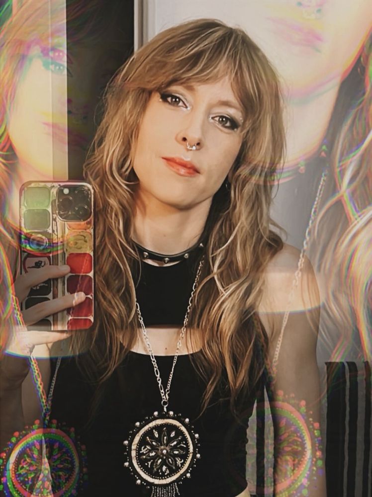 Femme presenting person smirks at the camera in a filtered selfie. They are wearing a beaded Métis pendant and have a blonde wolf cut. 