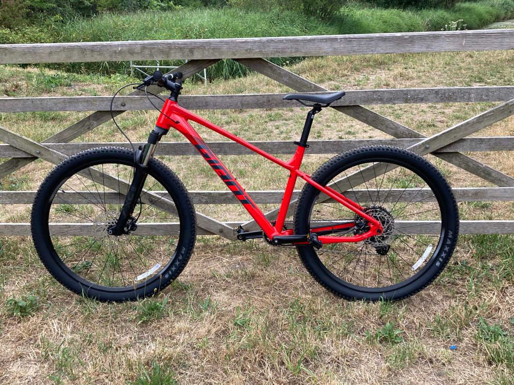 A photograph of a Giant “Talon 29 2” hard-tail bike propped against a wooden gate. The bike frame is bright red. The ground is covered with dry grass. There is more greenery in the background. 