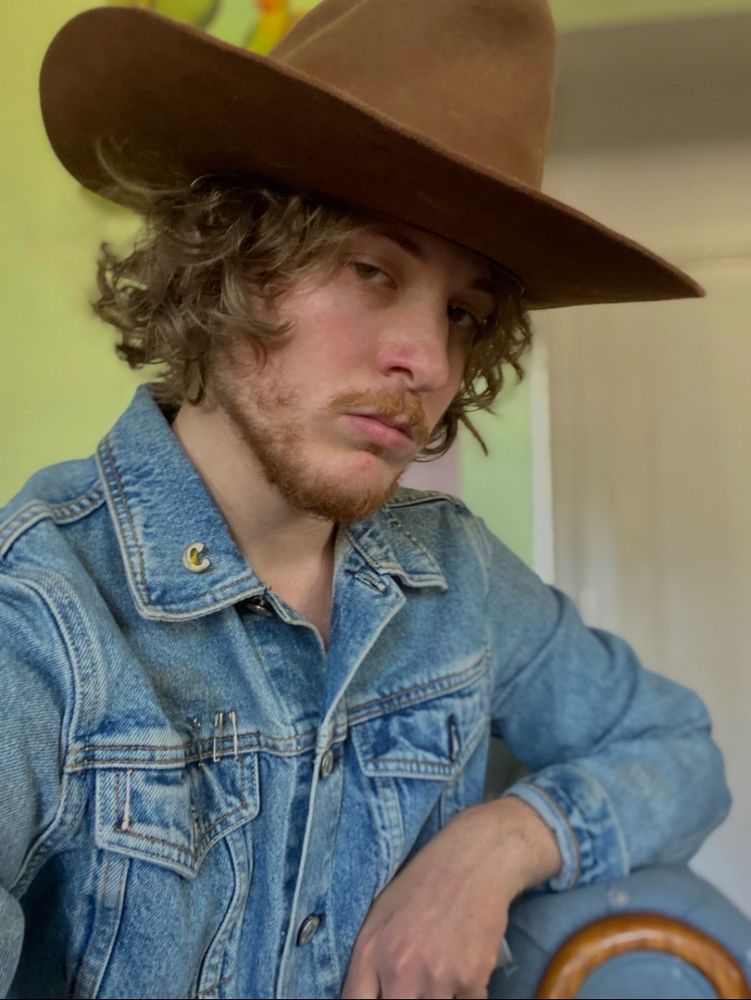 cher is sitting in his cabin with his elbow propped up on the arm rest of a blue couch while he looks at the camera. he is wearing a light blue jean jacket and a dark brown leather cowboy hat with his messy caramel colored hair popping out from beneath his hat. there is a golden charm with the letter “C” sewn into the collar of his jacket. his facial hair is scruffy. 