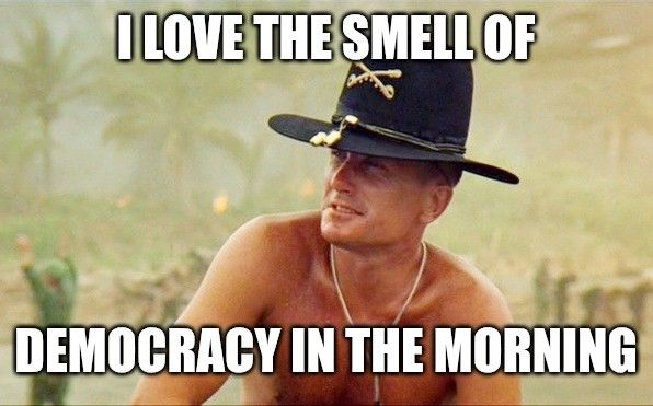 A still image of Robert Duvall in his role as Lt. William "Bill" Kilgore in the movie Apocalypse Now, shurtless and wearing a broad-brimmed cavalry hat with gold decorations. Superimposed text reads "I LOVE THE SMELL OF DEMOCRACY IN THE MORNING" 