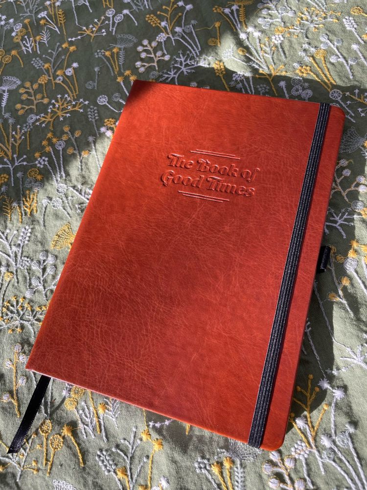 A brown leather bound book titled “The Book of Good Times” on top of a green, yellow, and white tablecloth.