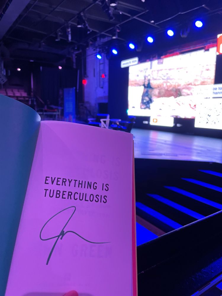A copy of Everything is Tuberculosis signed in green, in front of a stage with two chairs on it.