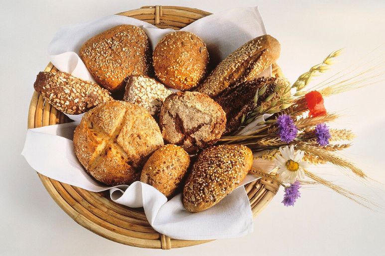 A wicker basket filled with a variety of seeded and whole grain bread rolls, placed on a white cloth napkin, accompanied by assorted wheat stalks and colorful wildflowers on the side.