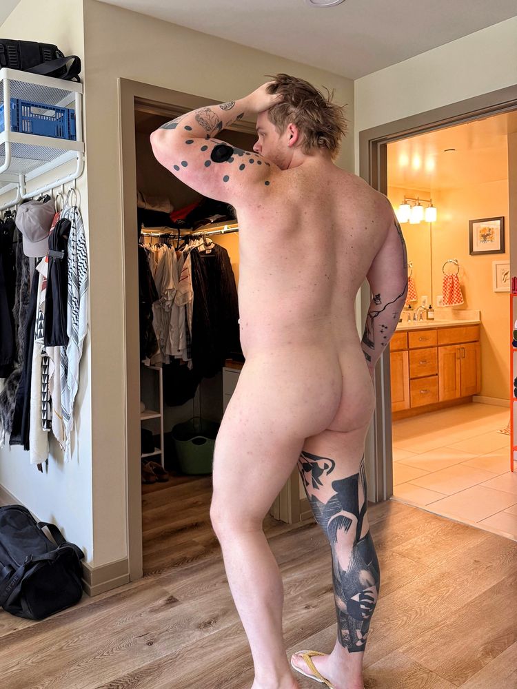 Me in my bedroom, I’m nude facing away from the camera, showing my large ass and semi-muscular back and legs. My arm is up tussling my hair.