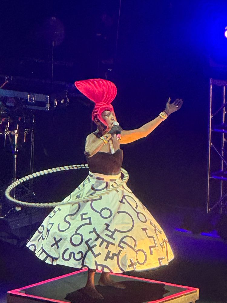 77-year-old Grace Jones hula hooping and singing into a microphone, wearing a black and white hoop skirt, black bustier and a dramatic red headpiece. 