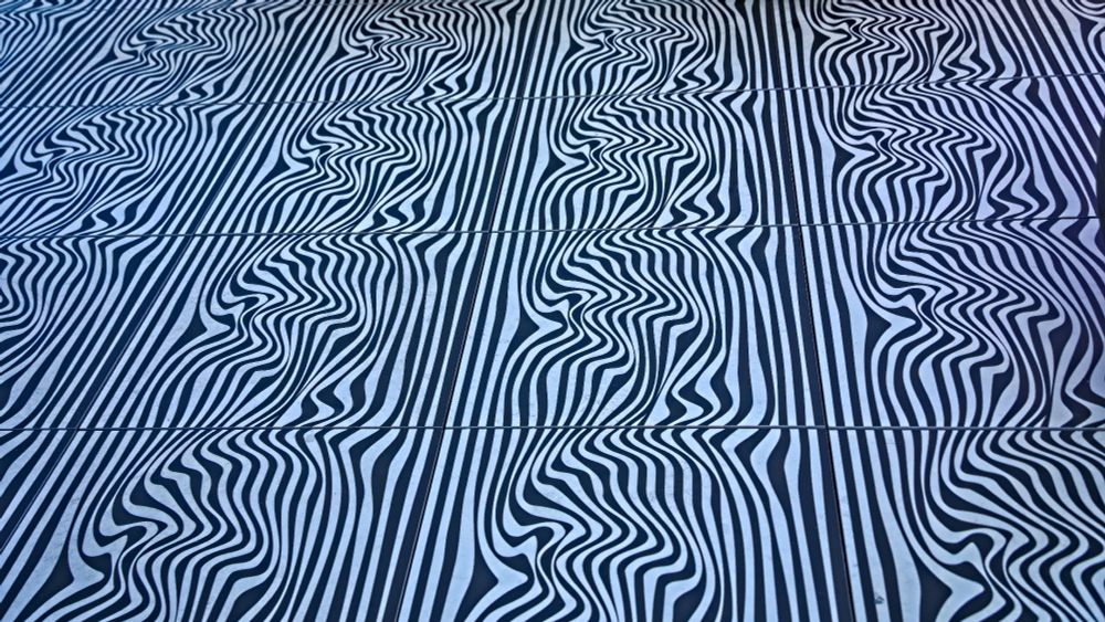 Dieses Foto zeigt eine abstrakte, grafische Oberfläche mit einem starken visuellen Effekt.

Das Bild besteht fast ausschließlich aus vertikalen Linien. Diese Linien sind abwechselnd schwarz und weiß. Das Muster ist nicht uniform: Die Linien erscheinen in regelmäßigen horizontalen Abständen periodisch verzerrt und wellenförmig (gewellt oder "flüssig"), während sie in den Zwischenräumen relativ gerade und parallel verlaufen. Diese abwechselnden Abschnitte erzeugen eine starke optische Täuschung von Bewegung, Tiefe und Wölbung.

Die Farben sind Schwarz und Weiß. 