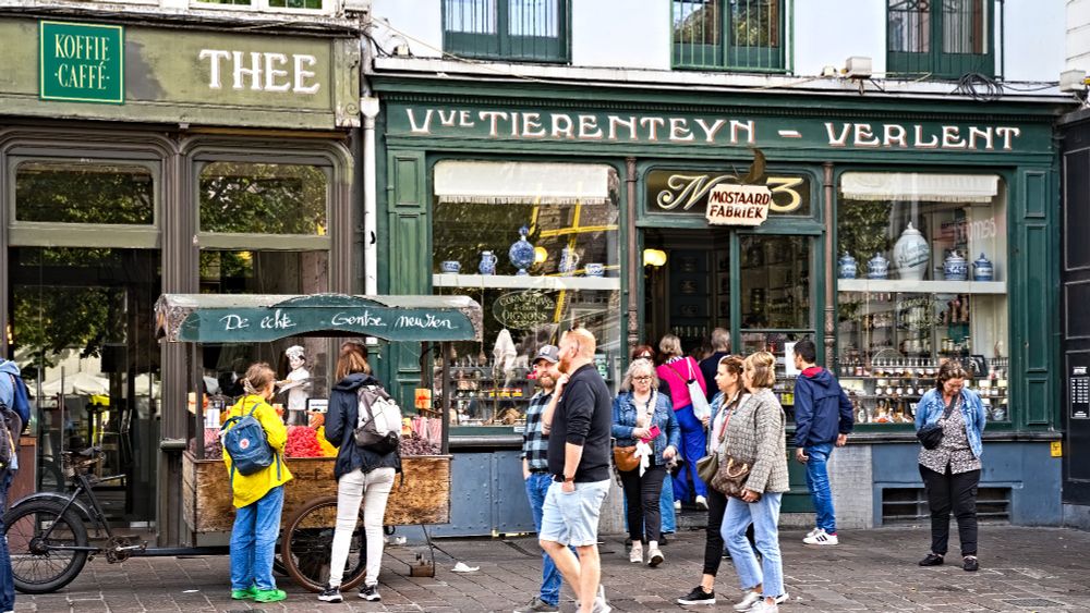 Das Foto zeigt eine belebte Szene in einer historischen europäischen Stadt, wobei zwei Geschäfte mit markanten, traditionellen Fassaden im Mittelpunkt stehen.

Zentrales Geschäft (Mitte und Rechts): Die zentrale Fassade ist in einem dunklen Flaschengrün gehalten.

Über dem großen Schaufenster steht in goldener Schrift der Name "VVE TIERENTEIN - VERLENT".

Darunter, in der Fenstermitte, ist kleiner der Schriftzug "MOSTAARD FABRIEK" (Senffabrik) zu lesen.

Das Schaufenster ist mit traditionellen Waren, blauen Keramikobjekten und Flaschen gefüllt.

Die Fassade links ist ebenfalls in einem dunklen, gedämpften Grün gehalten, teilweise abgesetzt mit weißer und goldener Schrift, die "KOFFIE CAFFÉ" und "THEE" (Tee) ankündigt.

Davor steht ein kleiner, mobiler Verkaufsstand mit der Aufschrift "de echte Gentse Meul...", der kleine Snacks anbietet.

Der Boden ist mit dunklem Kopfsteinpflaster belegt.

Eine Gruppe von Fußgängern dominiert den Mittelgrund.

Die Kleidung ist modern und lässig (Jeans, T-Shirts, Jacken).

Ein Mann und eine Frau mit Rucksäcken stehen links vor dem Verkaufsstand. Die Frau trägt eine leuchtend gelbe Regenjacke, was einen starken Farbakzent setzt.

Mehrere andere Personen, darunter Paare und Einzelpersonen, sind im Gespräch oder beim Flanieren zu sehen.