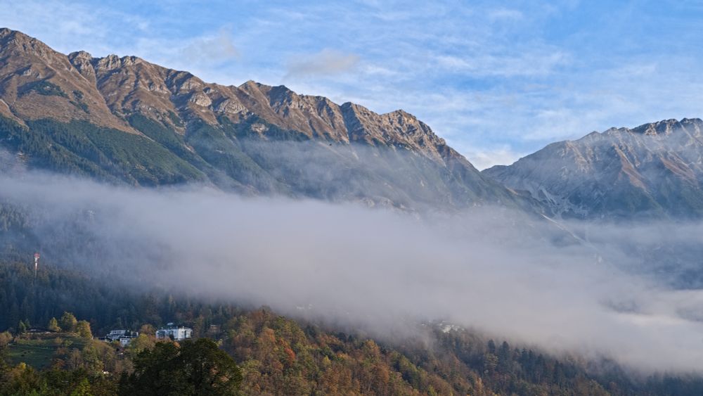 Das Foto zeigt eine beeindruckende Berglandschaft, die von einem dichten Nebelmeer umgeben ist. Die Berge ragen majestätisch aus dem Nebel heraus und sind in verschiedenen Brauntönen gefärbt, was auf eine herbstliche Jahreszeit hindeutet. Die Gipfel der Berge sind scharfkantig und wirken sehr imposant.
Der Nebel füllt die Täler und schafft eine mystische Atmosphäre, die den Blick auf die darunterliegenden Wälder und eventuell vorhandene Siedlungen teilweise verdeckt. Am unteren Rand des Bildes sind einige Bäume und Gebäude zu erkennen, die sich knapp über dem Nebel befinden.
Der Himmel darüber ist klar und blau mit einigen leichten Wolken.