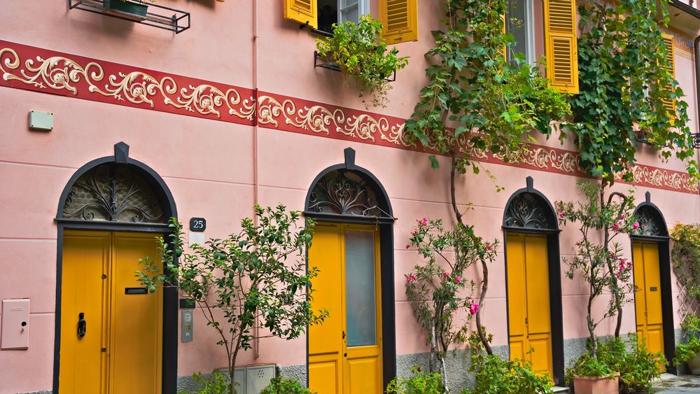 Das Foto zeigt eine Nahaufnahme eines farbenfrohen Gebäudes mit auffälligen architektonischen Details.


Die Wandfläche ist in einem sanften Rosa-Ton gestrichen.

Die Fassade wird durch ein breites, rotes Band horizontal unterteilt, das mit filigranen, cremefarbenen oder hellgelben Ornamenten und Arabesken verziert ist.

Die Erdgeschosszone wird von vier gelben Türen dominiert. Die Türen sind hoch und haben oben eine schwarze, halbkreisförmige Verzierung aus schmiedeeisernem Gitterwerk.

Die Fenster im oberen Stockwerk sind ebenfalls mit gelben Fensterläden versehen, die teilweise geöffnet sind.

Die Fassade ist lebhaft mit grüner Vegetation durchbrochen. Kleine Bäume oder Sträucher in Töpfen stehen vor den Türen.

Eine dichte Weinrebe oder Kletterpflanze rankt sich entlang der Wand und um die Fenster.

Aus den Fenstern hängen kleine Blumenkästen mit grünen Pflanzen.