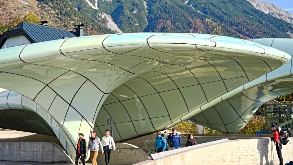 Das Foto zeigt eine futuristisch anmutende Station einer Bahn. Die auffälligste Struktur ist eine Dachkonstruktion aus Glas, die organisch gewellte Formen hat und einem Gletscher oder einer Wolke ähnelt. Die Oberfläche der Dachkonstruktion ist in einem hellen, leuchtenden Weiß-Grün-Ton gehalten und in ein Gittermuster aus dunklen Linien unterteilt. Unter der Überdachung sind mehrere Personen zu sehen, die in Alltagskleidung unterwegs sind, einige von ihnen mit Rucksäcken. Die gesamte Struktur steht in starkem Kontrast zu der dahinterliegenden Landschaft: majestätische, bewaldete Berge mit felsigen Gipfeln unter einem blauen Himmel. Am rechten Bildrand ist eine Person mit Wanderstock und rotem Rucksack zu erkennen. 
