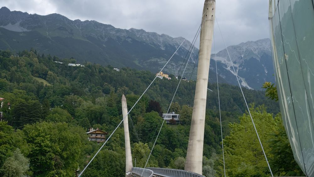 Das Foto zeigt eine Aussicht von der Station der Hungerburgbahn im Tal. Die Station und die Strecke sind durch große, schräge Betonpfeiler und Stahlseile stabilisiert, die eine futuristische und elegante Architektur schaffen. Im Hintergrund sieht man eine üppige, grüne Berglandschaft mit dichten Wäldern und vereinzelten Gebäuden, die sich harmonisch in die Natur einfügen. Die Berge im Hintergrund sind teilweise bewaldet und zeigen schroffe Felsformationen, die in den Himmel ragen. Der Himmel ist leicht bewölkt, was der Szene eine ruhige und friedliche Atmosphäre verleiht. Die Perspektive des Fotos betont die Höhe und Weite der Landschaft und vermittelt ein Gefühl von Freiheit und Weite. Die Kombination aus moderner Architektur und Natur schafft einen Kontrast.