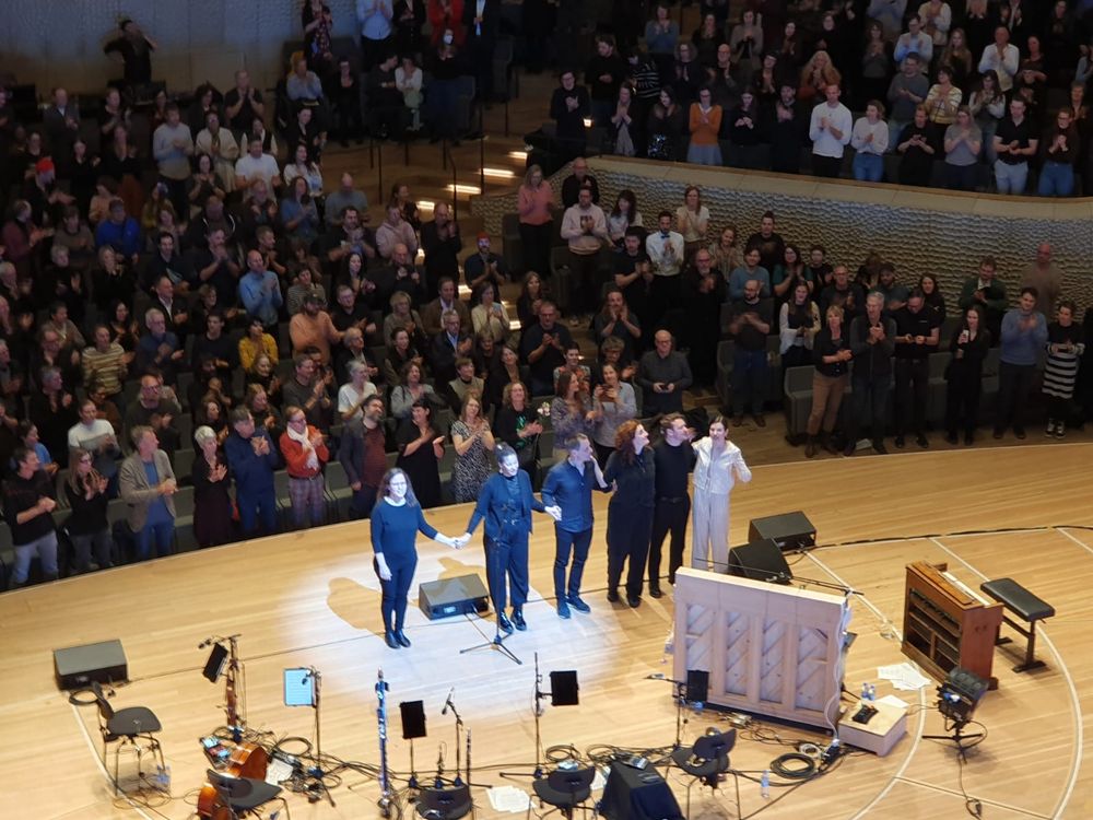 Abschlussapplaus. Die sechs Musiker:innen stehen Arm in Arm auf der Bühne. Das Publikum gibt Standing Ovation