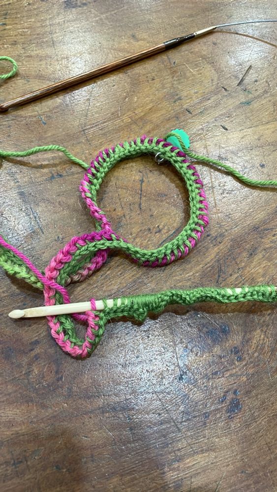 A coil of pink and green crochet stitches lie on a wooden surface with a wooden crochet hook attached.