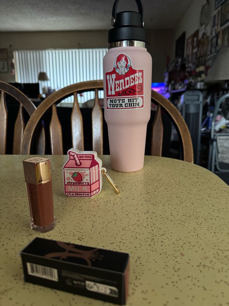 A light pink water bottle with the Wendy’s fast food label changed to read, Wendeez Nuts Hit Your Chin, an AirPods case in the shape of a strawberry milk carton, and a brown Anastasia Beverly Hills lip gloss on our kitchen table.