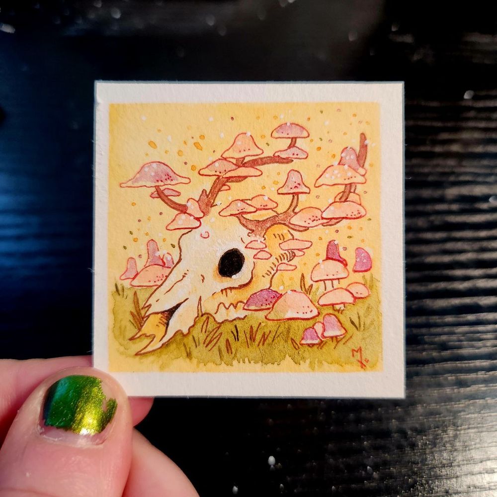 photo of me holding the mini painting. painting is a deer skull with mushrooms growing on the brain cavity, antlers, and surrounding grass. spores dotting the empty space around the focal point.