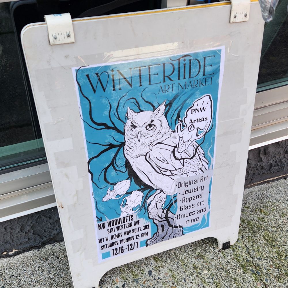 A sign with a graphic of an owl by artist Stef Azevedo that reads "Wintertide Art Market - NW Worklofts 3131 Western Ave 107 W Denny Way Suite 303 Saturday and Sunday 12-6 12/6-12/7 - PNW Artists - Original Art Jewelry Apparel Glass Art Knives and more