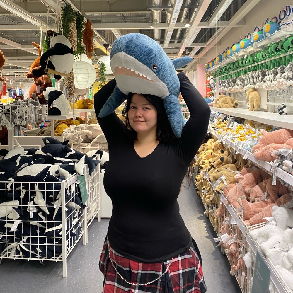 AngelNaomi standing straight holding cute shark plush blahaj while standing in ikea. AngelNaomi is wearing a black shirt and plaid skirt, while the background shows rows of other plushies.