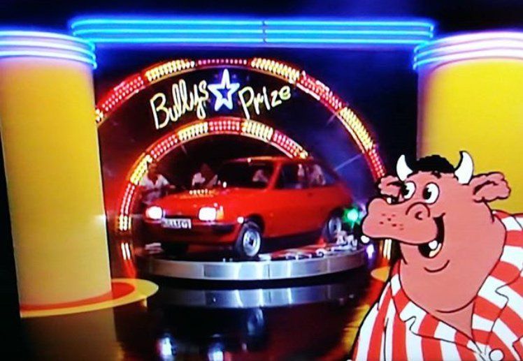 A scene from the world's greatest television program, Bullseye with Jim Bowen, showing the star prize which is a red car with a registration plate that reads Bullseye. Its headlights are on for some reason. It is on a circular platform designed to resemble a gigantic dartboard, and several poor souls from backstage have been wrangled to push it out onto the main stage. A cartoon rendition of the show's mascot, Bully, is in the bottom right corner of the screen. I'm not sure if the contestants won the car or not, but as they have not yet been herded towards it by Jim Bowen I find it unlikely.