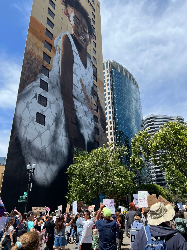A large group of protesters marching beneath a huge mural of a Black girl called “No Hunger” in Oakland, CA