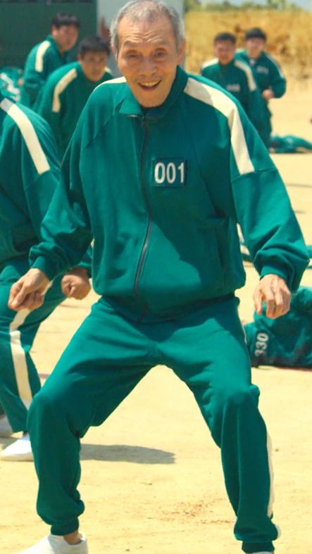 screenshot from squid game s01e01 featuring a grinning old man participating in the games, there are scared participants and laying bodies in the background