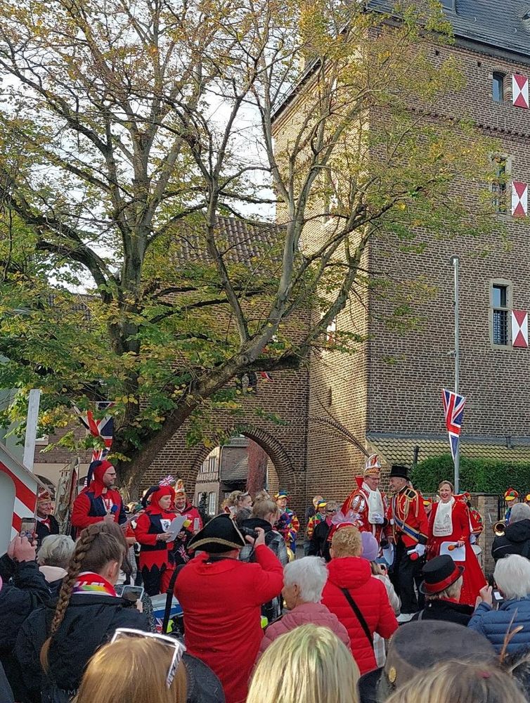 festliche Straßenszene vor einem historischen Gebäude mit Fachwerk- und Backsteinarchitektur. Eine große Gruppe von Menschen, viele in bunten oder traditionellen Kostümen, hat sich versammelt. Im Mittelpunkt stehen Personen in rot-weißen Karnevalskostümen, darunter ein Prinzenpaar und andere Teilnehmer in närrischer Kleidung.