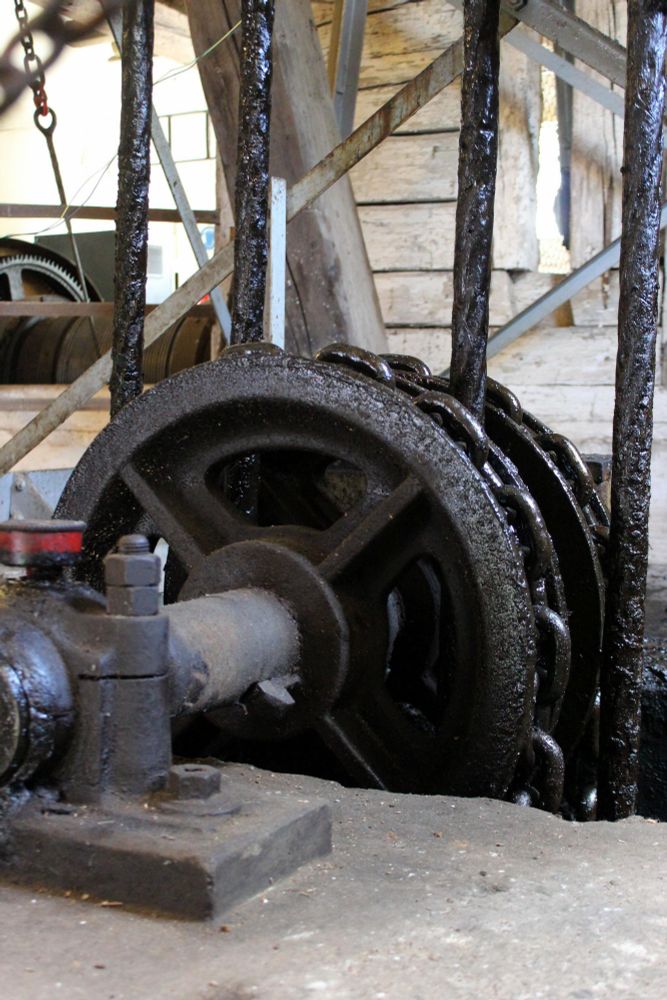 Nahaufnahme eines alten, stark geölten Zahnrads mit Kettenantrieb. Die Umgebung zeigt metallene Streben und verwittertes Holz