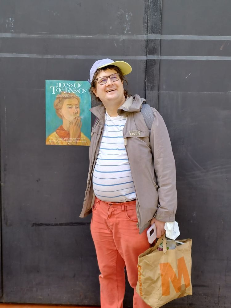 Moi avec une casquette me tenant devant une affiche de l'exposition "House of Tove Jansson" qui représente un autoportrait de Tove en train de fumer. Je souris
