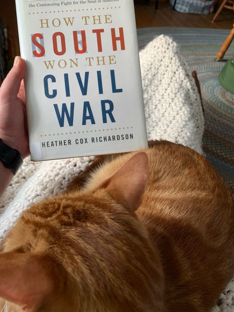 An orange cat on OP’s lap, OP is holding a library copy of Heather Cox Richardson’s book “How the South Won the Civil War”. 