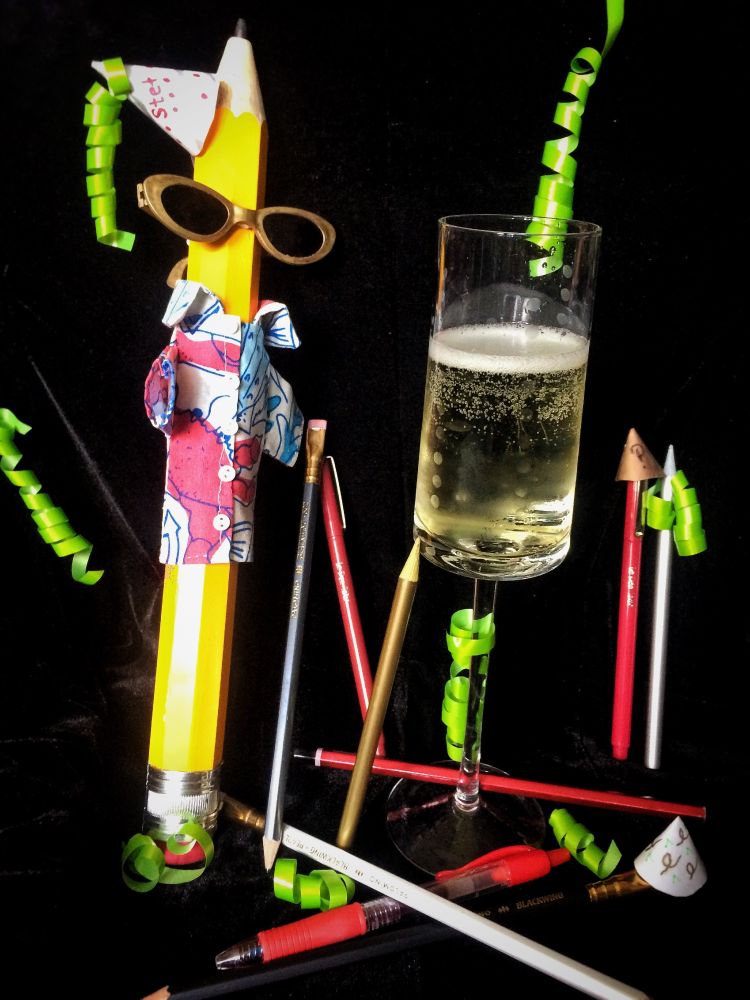 Giant Pencil wearing tropical shirt and sunglasses with a tiny party hat, surrounded by various writing implements, a glass of champagne, tiny editing themed party hats, and neon green ribbon curls