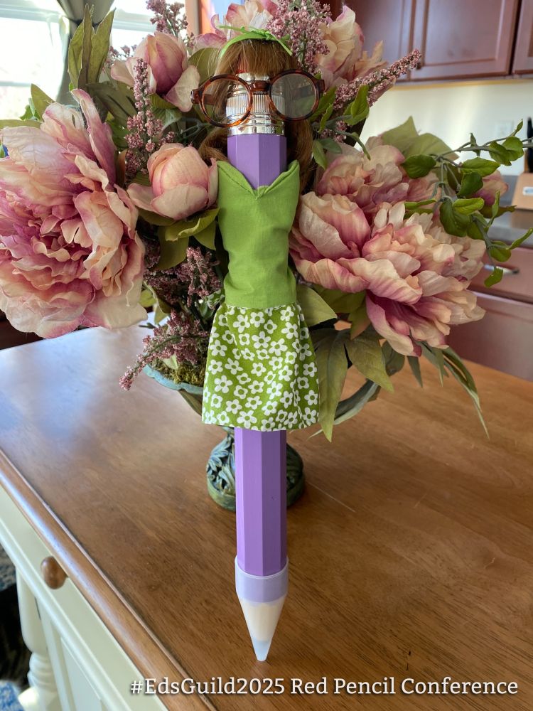 Julie Willson's "Pencelope," a giant purple pencil wearing a green floral dress with round glasses and a tiny wig, posing in front of a bouquet of flowers while watching the conference online.
