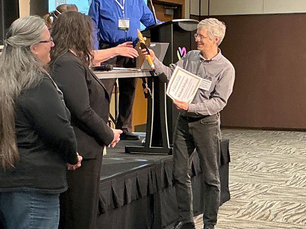 Michael Schuler accepting the Guild Award of Excellence and smiling while holding Guild mascot Giant Pencil, which is wearing a tiny tuxedo.