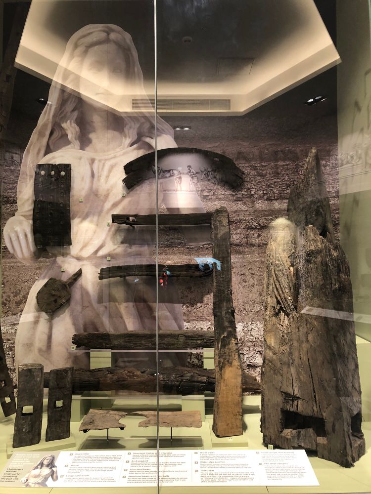 Another display case in the Wood gallery at the Chesterholm Museum at Vindolanda - with many more wooden artefacts including parts of large wooden vessels, and several smaller utensils- with a beautiful backdrop of a Roman women illustrated on the rear wall of the case. Photo copyright Coralie Mills 2024.