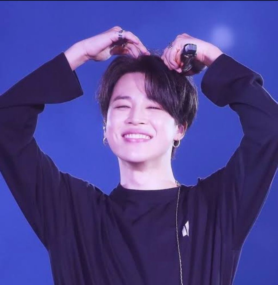 Jimin is smiling while holding a microphone and doing a heart pose over his head.