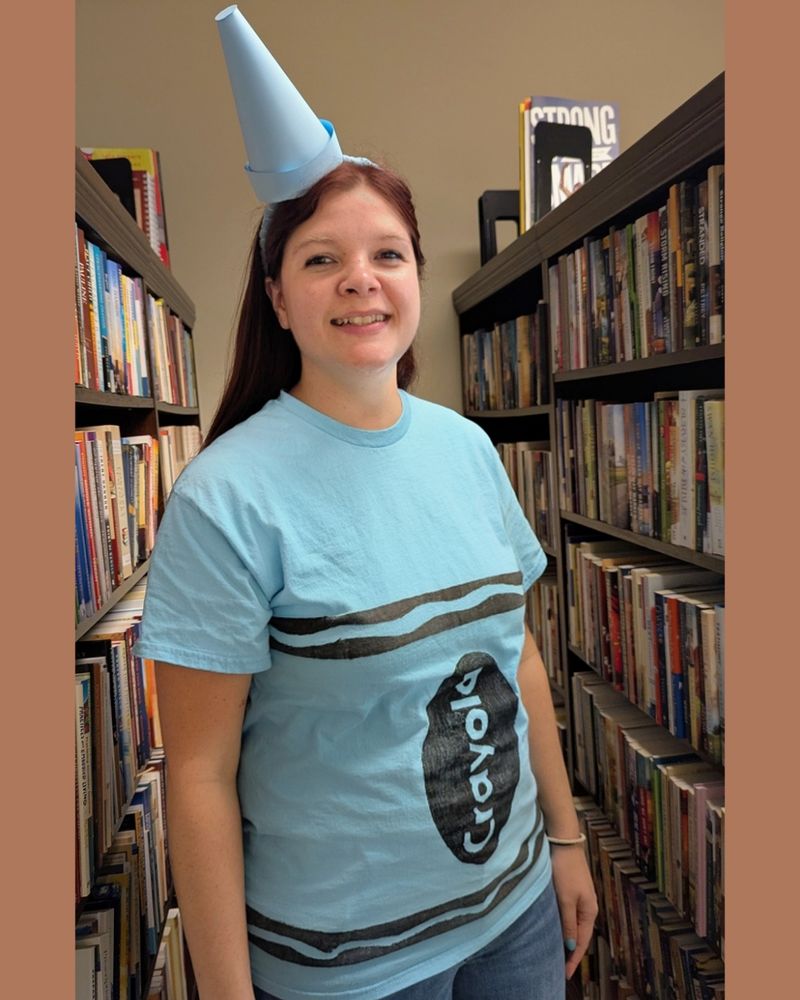 A woman wears a light blue shirt crafted to look like a Crayola crayon. A crayon tip-shaped hat is perched askew on her head. 