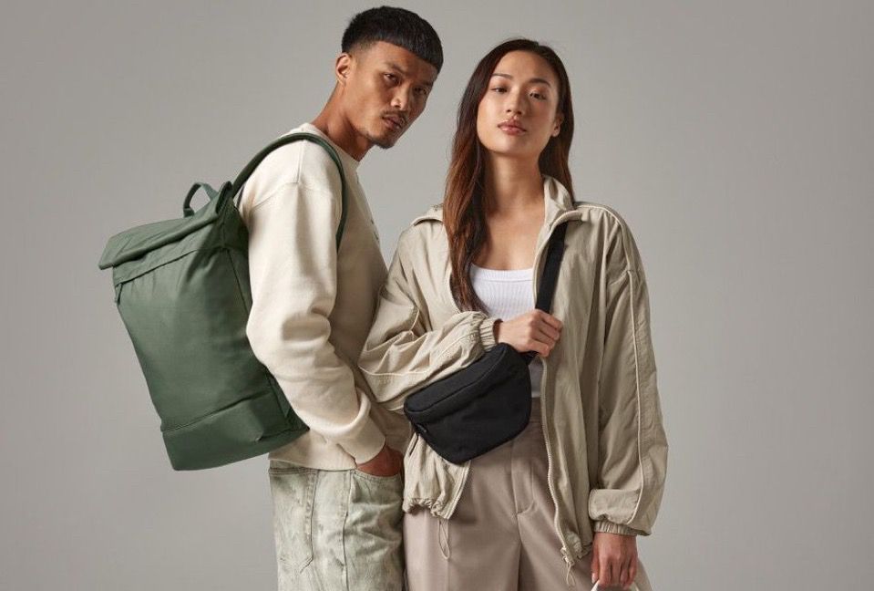 A male and female model with BagBase's Simplicity Roll-Top Backpack. Made from a 100% recycled 600D polyester fabric, this water-repellent backpack has an ergonomic design with padded tech protection and a signature roll-top closure.