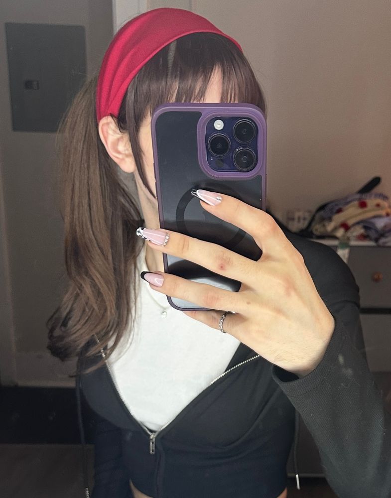 selfie of a trans girl sitting in front of a makeup vanity in a black hoodie with her hair in a ponytail and headband with cowprint accented nails