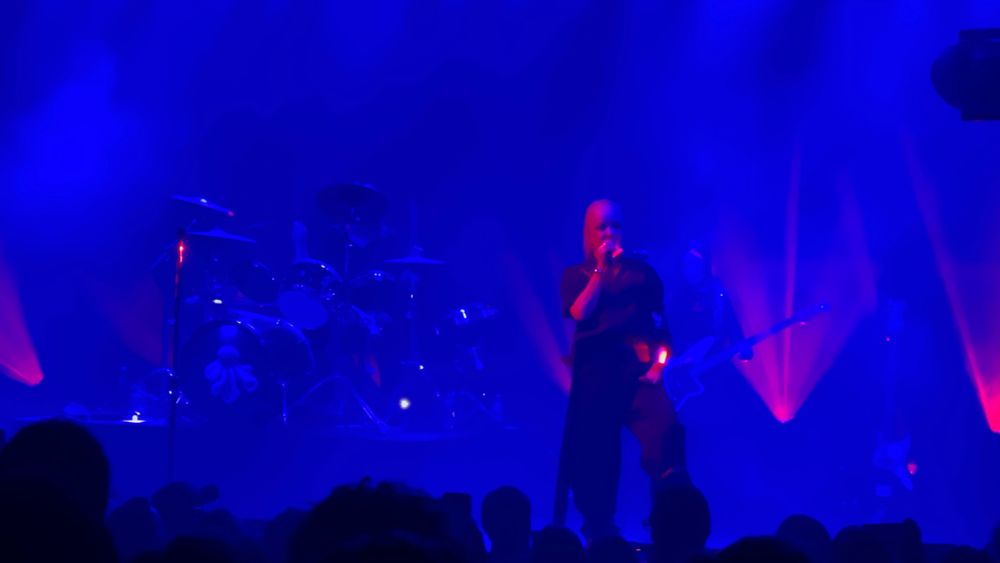A concert stage with the band Garbage performing, and heads and hands in the foreground. The lighting is blue with some red in the background