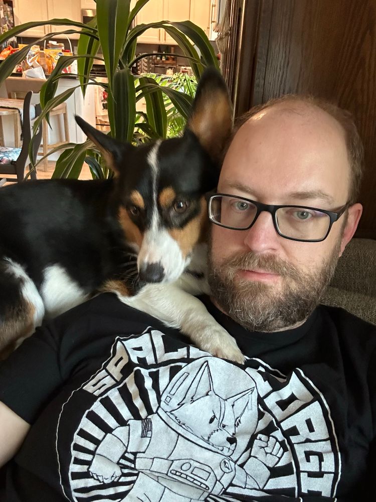 Frankie the tricolor corgi chilling on the back of the couch and resting his paw on Vidmaster’s shoulder. Vidmaster is a balding middle aged white dude with glasses and a beard, and he is wearing a black Space Corgi t-shirt.