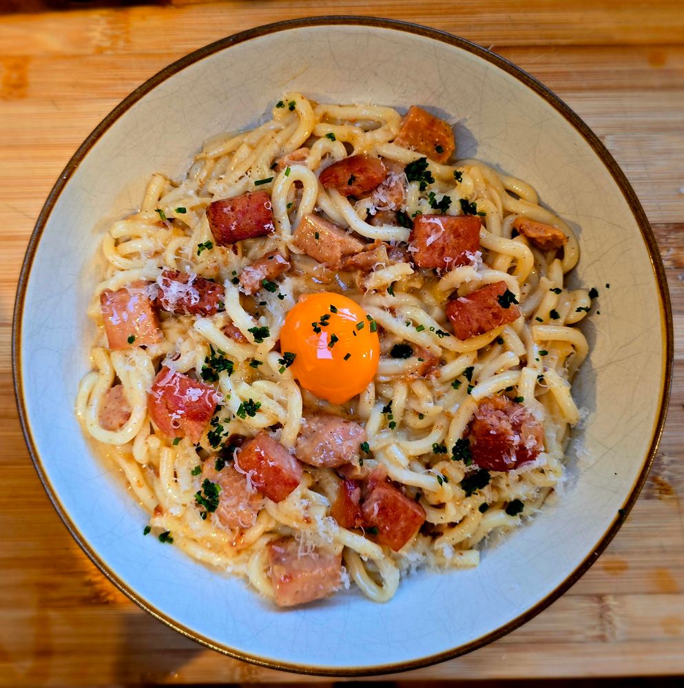 A large bowl of udon noodles in a creamy sauce with large golden chunks of spam and a single vibrant orange egg yolk in the middle. 