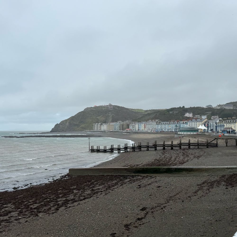Aberystwyth north beach, a gravelly beach in a dark grey, rainy setting. The bay is lined with colourful terraced buildings.