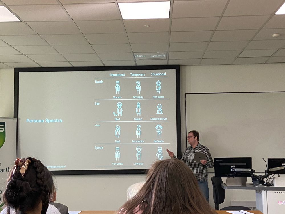 Photo of the poster, Felix at the front of a university lecture room, he is pointing at the content on the screen, which is an example of a persona spectrum, illustrating how a permanent, temporary or situational loss of some of our senses impact different people.