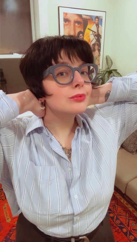 FGB highlighting her new micro French bob with bangs. They have dark almost black-brown hair and are wearing blue glasses stands indoors. They're wearing a light blue, vertically striped button-down shirt tucked into high-waisted dark trousers. Behind them their spare bedroom living space. There is a beige couch with a dog-shaped pillow, a bright red patterned red rug, a leafy potted plant on a small wooden stand, and a framed vintage movie poster featuring dramatic portraits and bold colors. In this pic FGB is lightly fluffing the bottom of her short bob that she is very happy with rn.