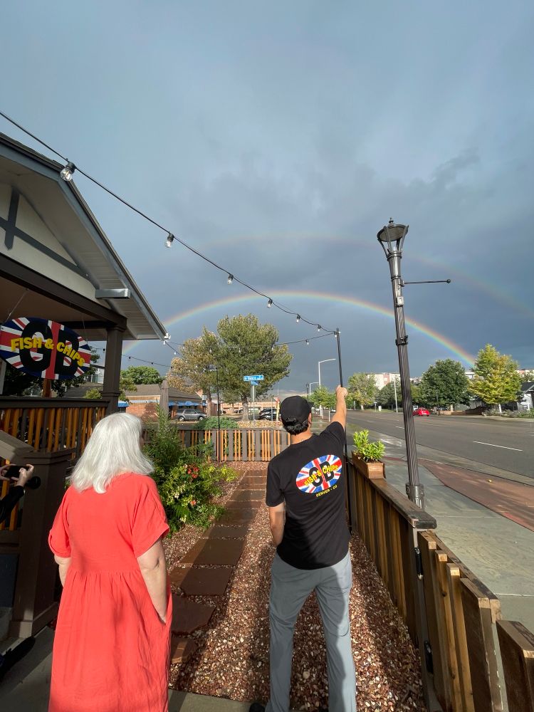 A woman in a red dress and a man in a staff shirt both stand with their backs to the camera facing a double rainbow, the man is pointing 