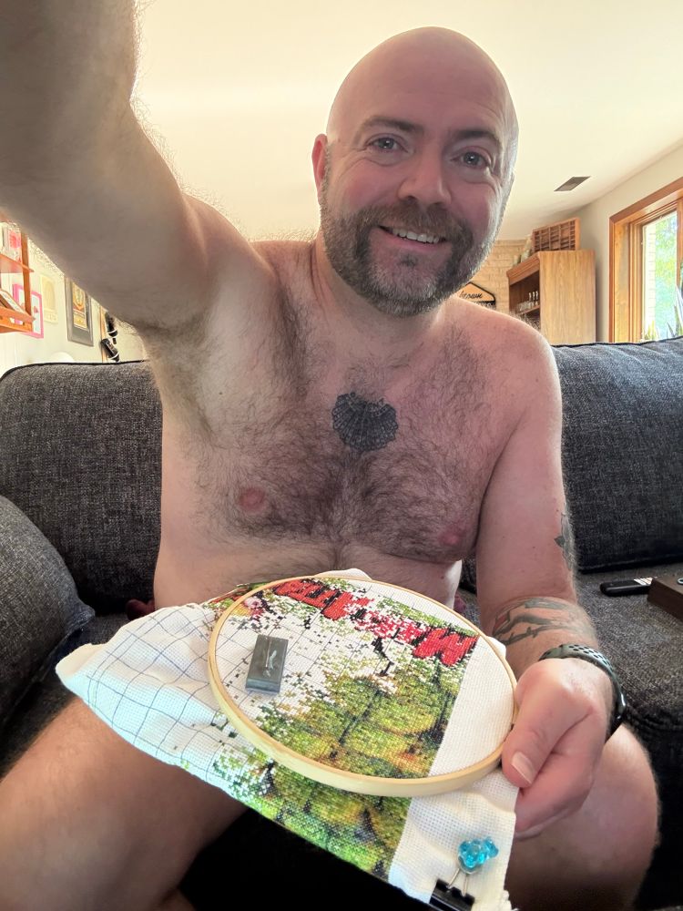 Cole sitting on a couch with an embroidery hoop with a cross stitch project in it. The project is strategically placed to question whether or not he is wearing pants.
