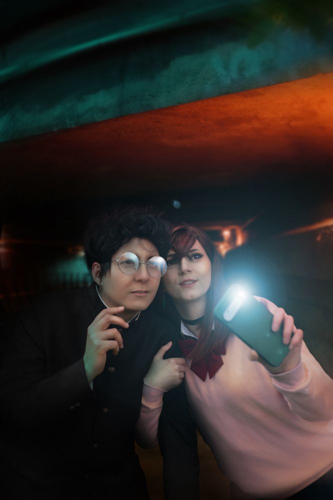 Cosplayer as Okarun and Momo from Dandadan, Momo holding a phone with flash up, both looking skeptical, a red/turquoise lighted underpath in the background