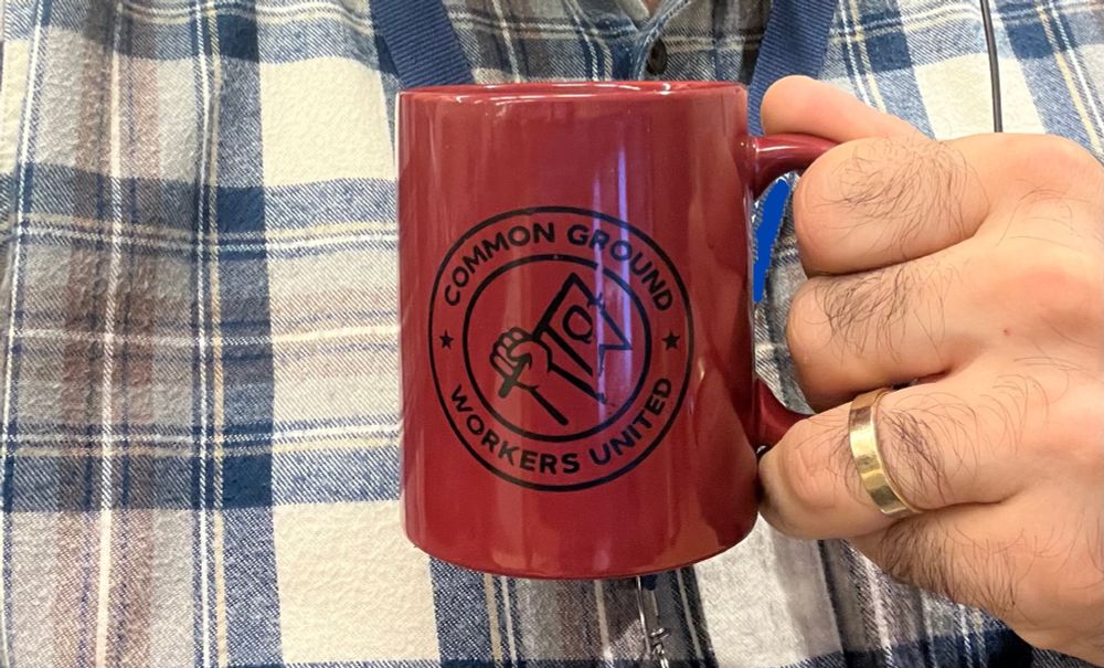 A Common Ground Workers United coffee mug in front of a blue white and brown flannel shirt 
